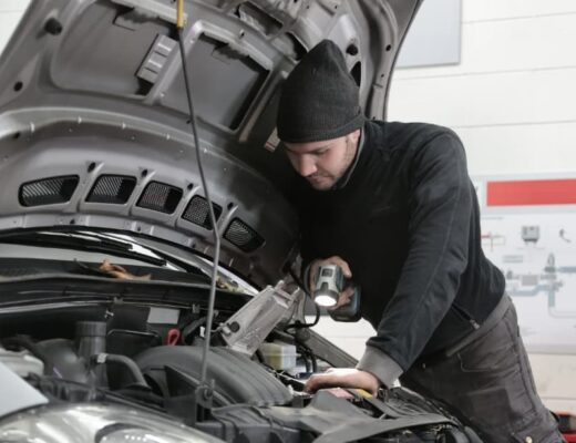 a man in a hat looking under the open hood of a car with a flashlight in his hand