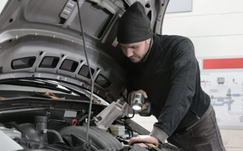 a man in a hat looking under the open hood of a car with a flashlight in his hand